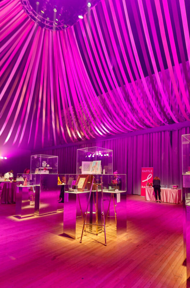Elegant event venue space with a purple lighting scheme, draped ceiling decorations, and display tables with exhibits.