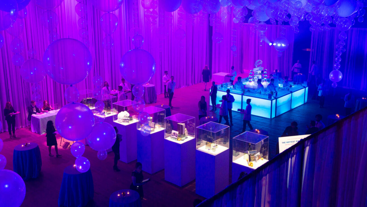 An indoor event venue space illuminated with purple lighting, adorned with large balloons, and attendees mingling near exhibits.