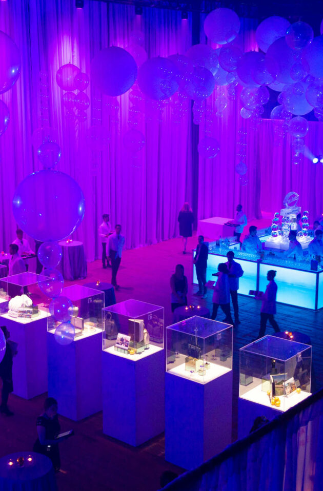 An indoor event venue space illuminated with purple lighting, adorned with large balloons, and attendees mingling near exhibits.