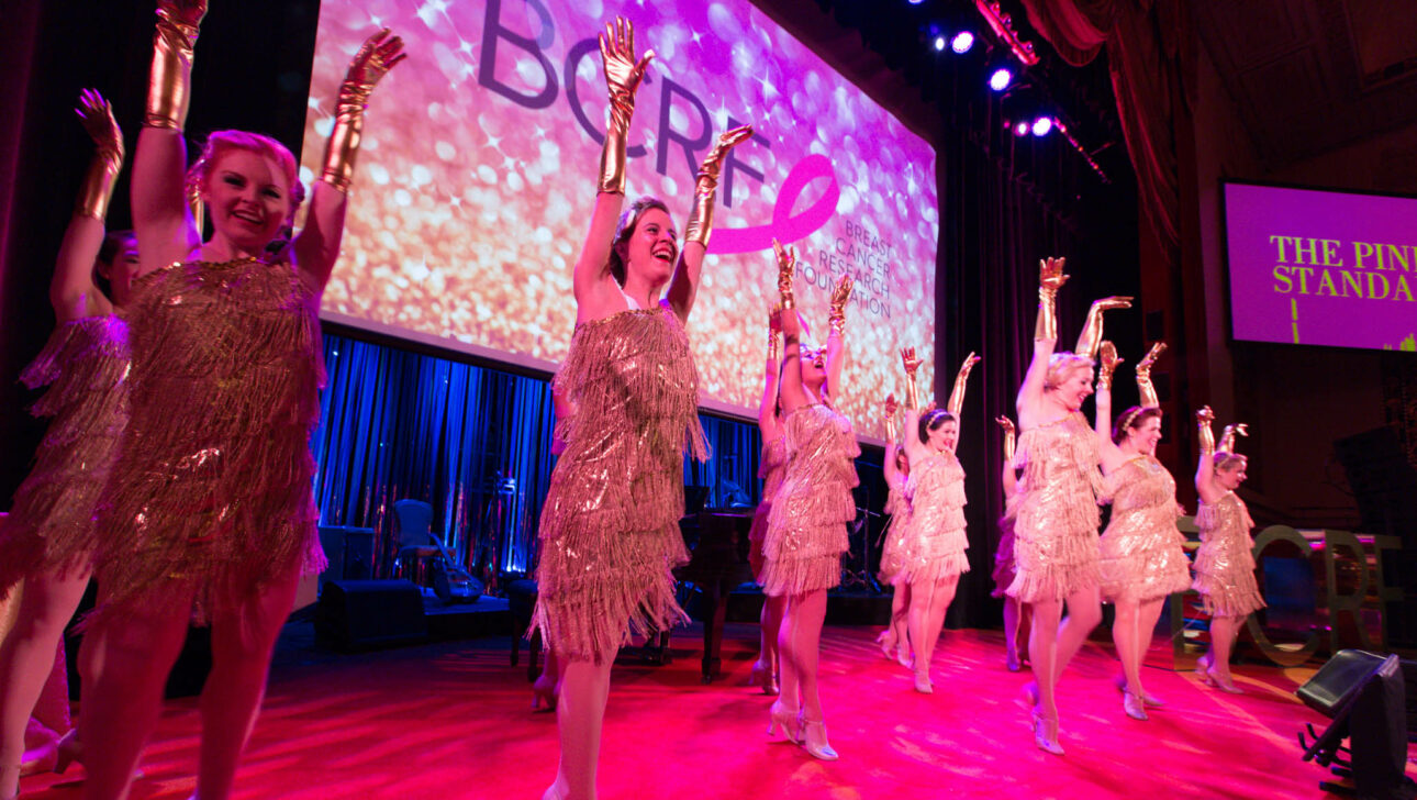 Performers in sparkling costumes dancing on stage at an event with the word "broad" projected in the background, organized by a renowned event planning company.