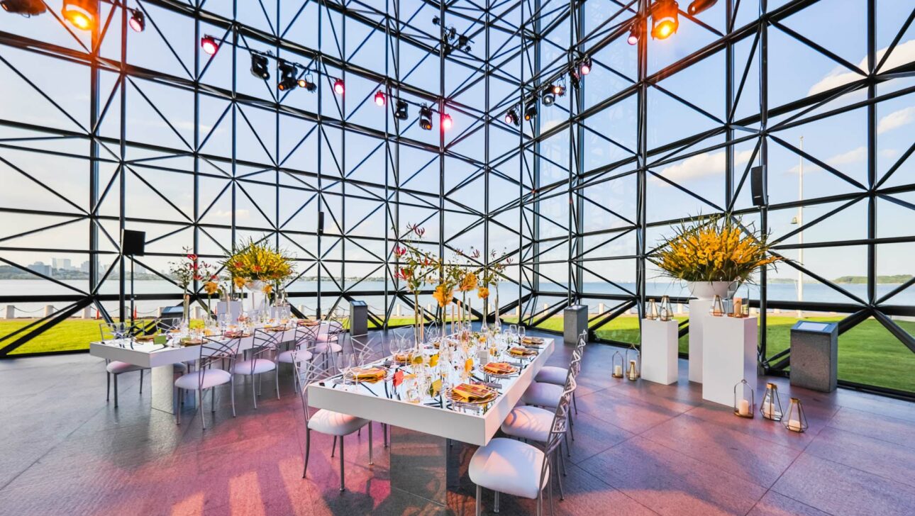 A modern event venue space with a geometric glass facade, elegantly set dining tables, and a view of the water and skyline.