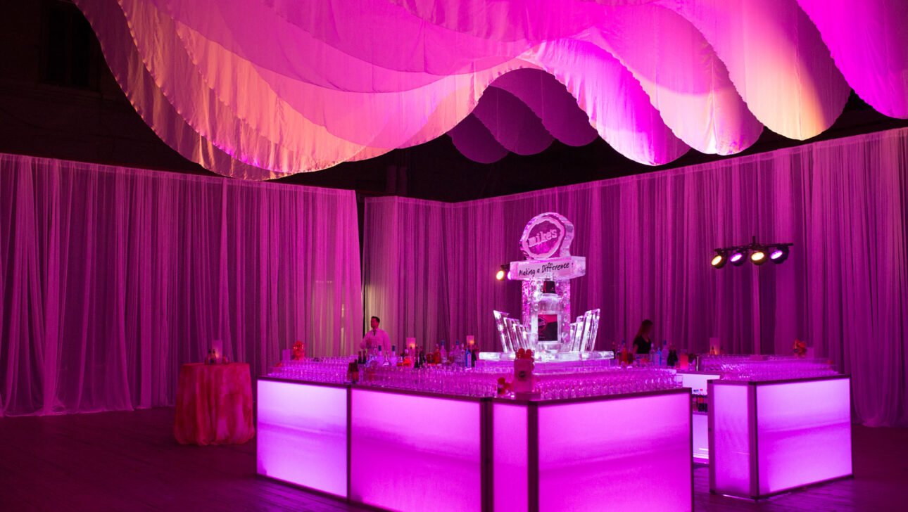 Elegant event setup by professional event planners with a lit bar and ice sculpture under pink lighting.