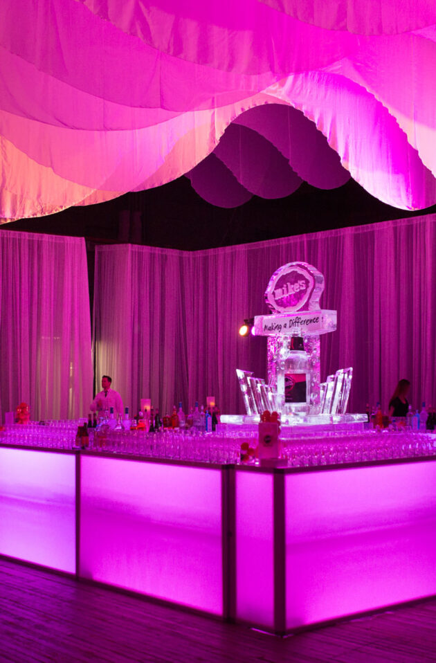 Elegant event setup by professional event planners with a lit bar and ice sculpture under pink lighting.
