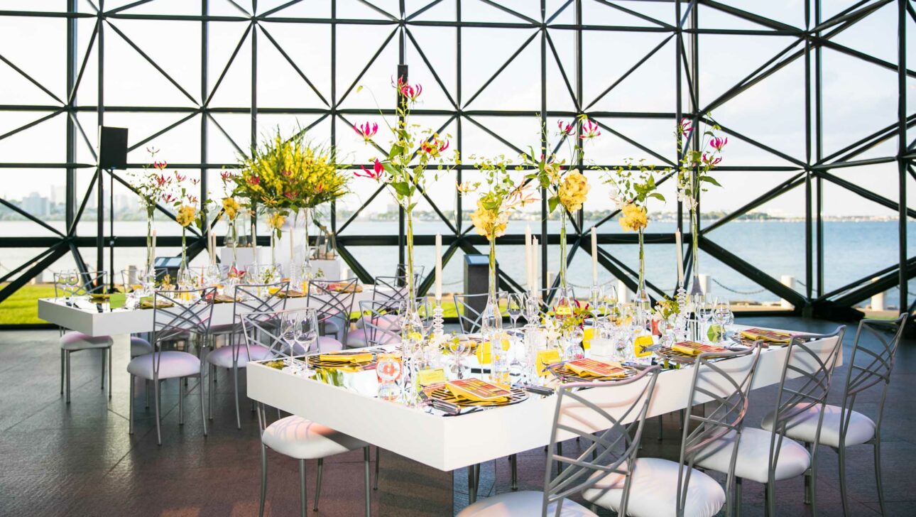 Elegant dining setup with floral centerpieces in a modern event venue overlooking the waterfront.