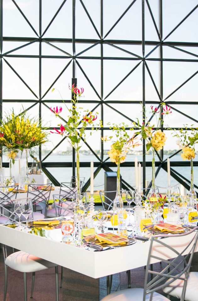 Elegant dining setup with floral centerpieces in a modern event venue overlooking the waterfront.