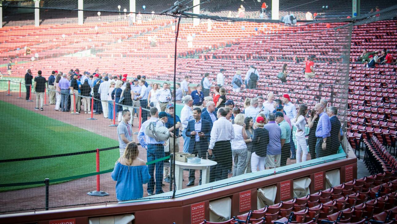 Group of people mingling in a baseball stadium, an event venue space, with empty red seats in the foreground.