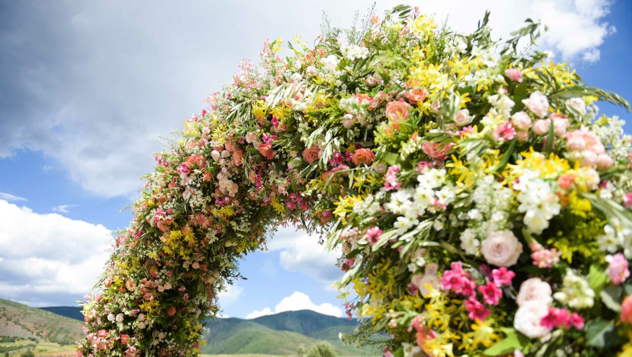 Floral archway, designed by top event planners, with a variety of colorful flowers, set against the backdrop of a mountainous landscape and a partly cloudy sky.
