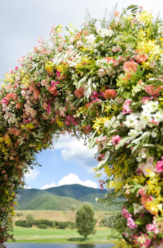 Floral archway, designed by top event planners, with a variety of colorful flowers, set against the backdrop of a mountainous landscape and a partly cloudy sky.