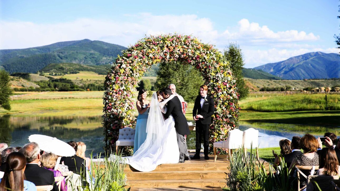 A wedding ceremony organized by an event planning company, taking place outdoors with a floral arch, a couple at the altar, and guests seated facing a scenic mountain backdrop.