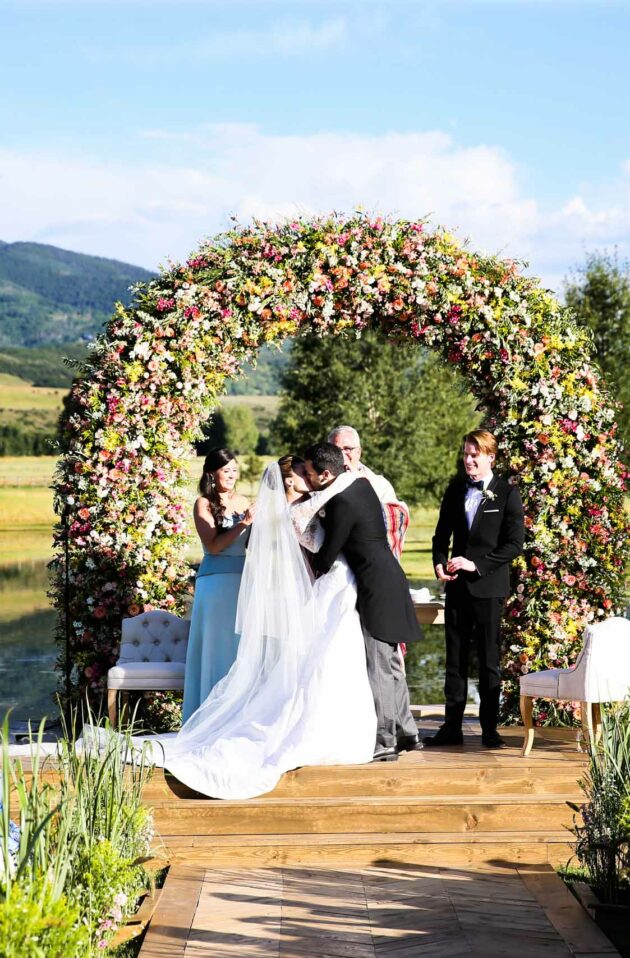 A wedding ceremony organized by an event planning company, taking place outdoors with a floral arch, a couple at the altar, and guests seated facing a scenic mountain backdrop.