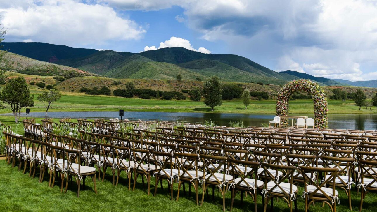 Outdoor wedding venue set up by professional event planners with rows of chairs and a floral arch near a tranquil lake with mountains in the background.