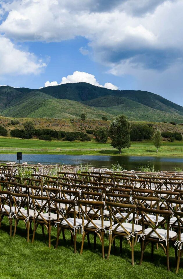 Outdoor wedding venue set up by professional event planners with rows of chairs and a floral arch near a tranquil lake with mountains in the background.