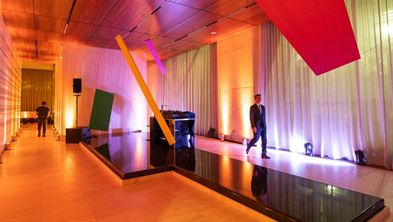 Modern event venue space with colorful lighting and abstract art installations, featuring a reflective water feature and individuals walking through the area.
