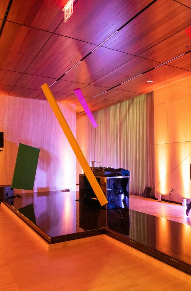 Modern event venue space with colorful lighting and abstract art installations, featuring a reflective water feature and individuals walking through the area.