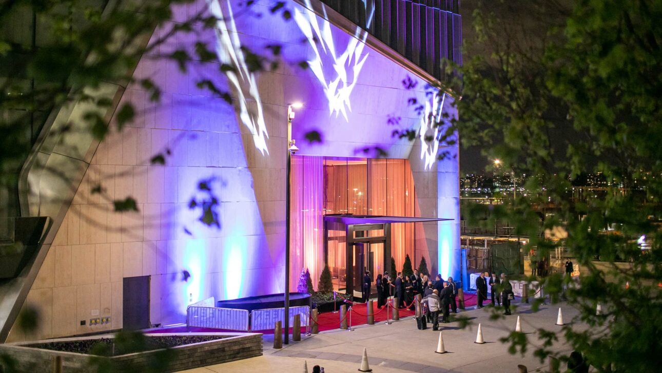 An illuminated event venue entrance with people waiting in line at night, featuring a red carpet and decorative lights.