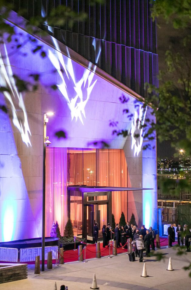 An illuminated event venue entrance with people waiting in line at night, featuring a red carpet and decorative lights.