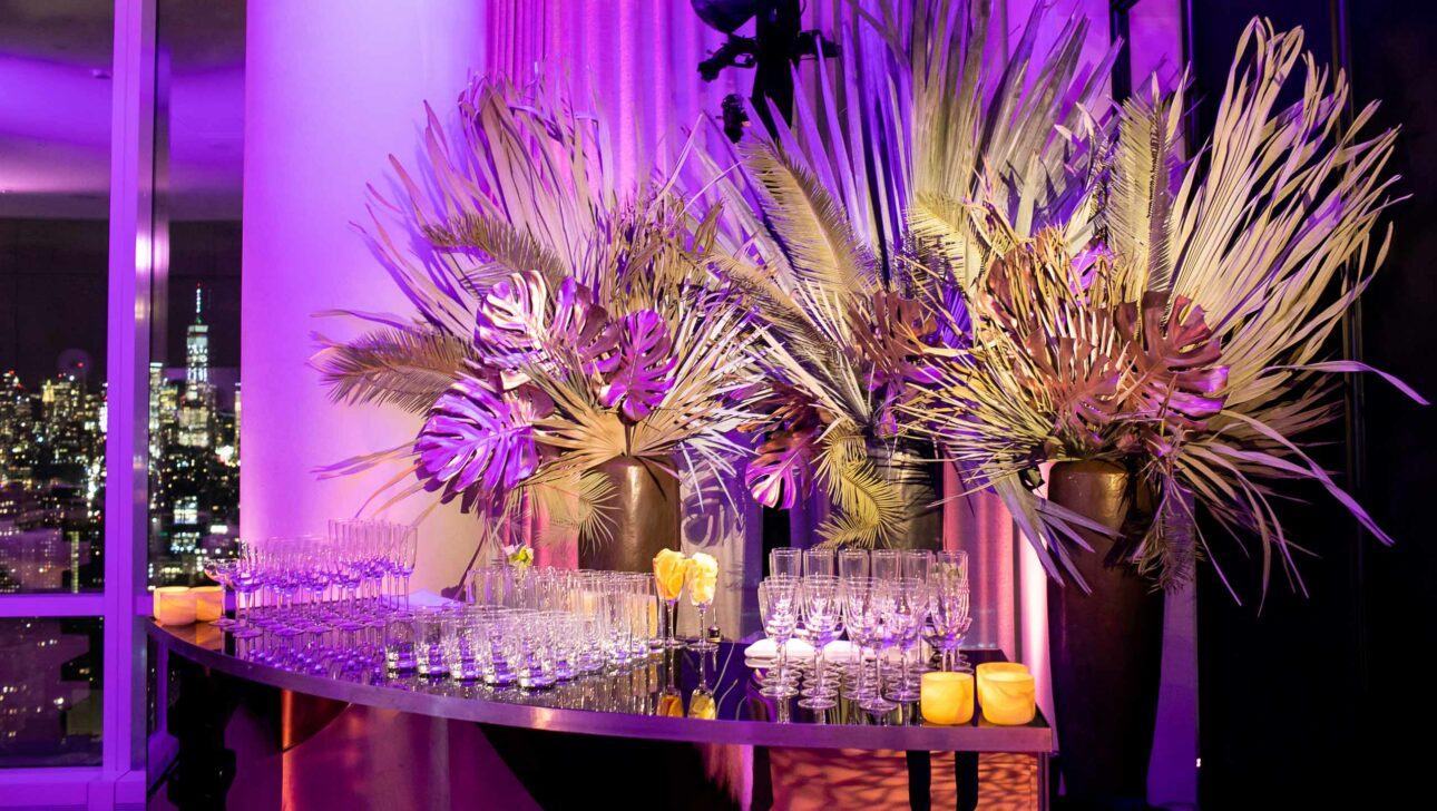 Elegant event setup by an esteemed event planning company, with an array of glasses on a table adorned with tropical leaves and flowers, featuring city skyline views at dusk.