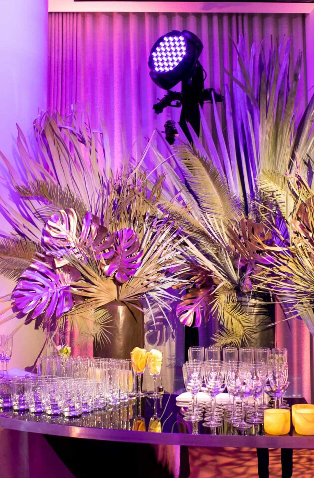 Elegant event setup by an esteemed event planning company, with an array of glasses on a table adorned with tropical leaves and flowers, featuring city skyline views at dusk.