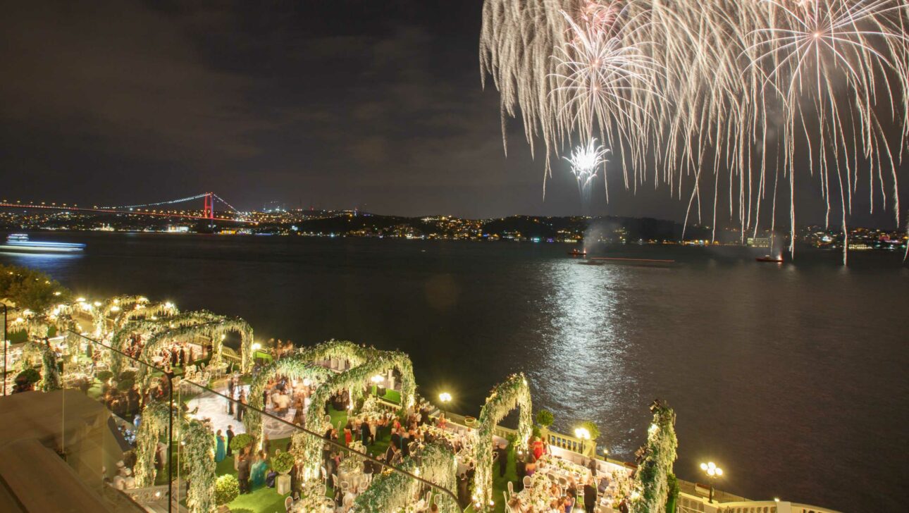 Fireworks illuminate the night sky above a river, with a festive outdoor gathering organized by an event planning company visible and a bridge in the background.