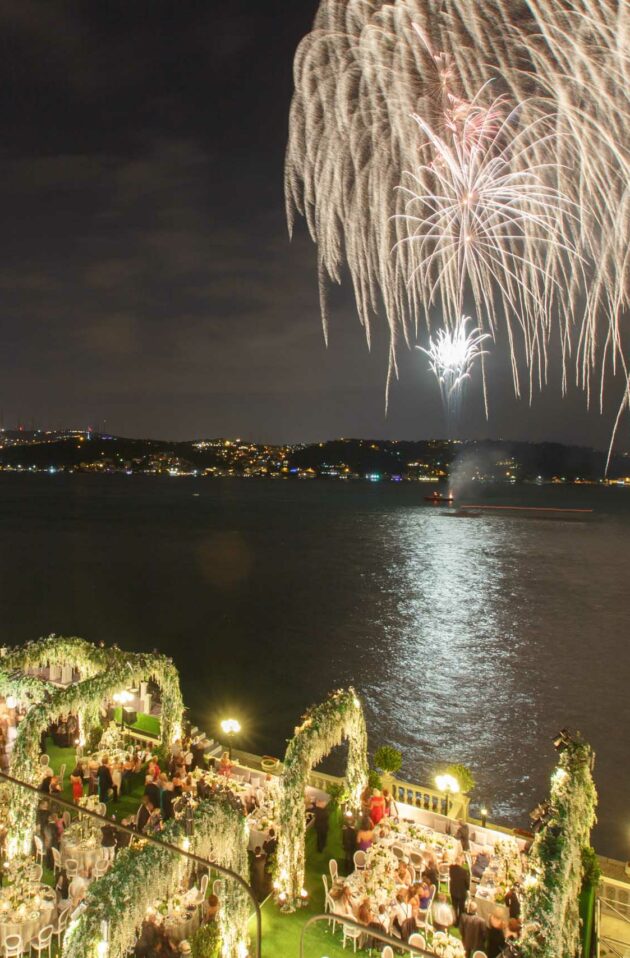 Fireworks illuminate the night sky above a river, with a festive outdoor gathering organized by an event planning company visible and a bridge in the background.