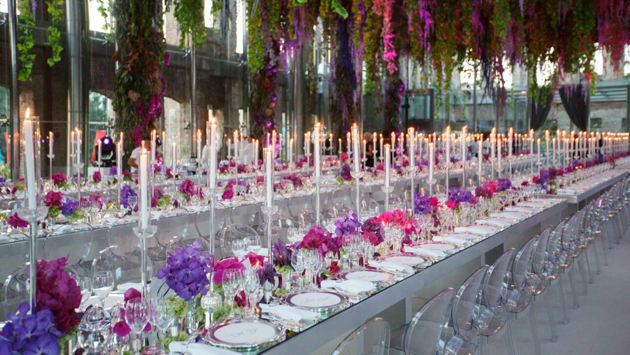 Elegant banquet setup by event planners with long tables, candlelight, and hanging purple floral arrangements.
