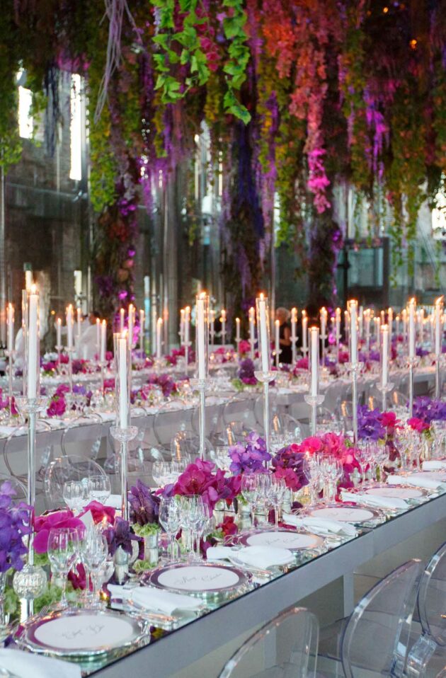 Elegant banquet setup by event planners with long tables, candlelight, and hanging purple floral arrangements.