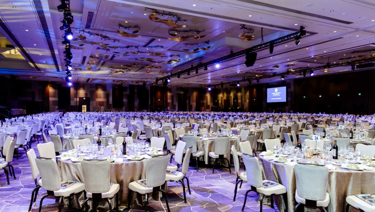 Banquet hall set up for a formal event by an event planning company, with numerous dining tables and a stage with a projection screen.