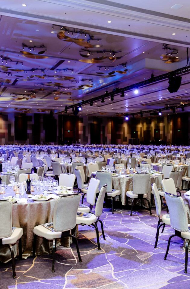 Banquet hall set up for a formal event by an event planning company, with numerous dining tables and a stage with a projection screen.